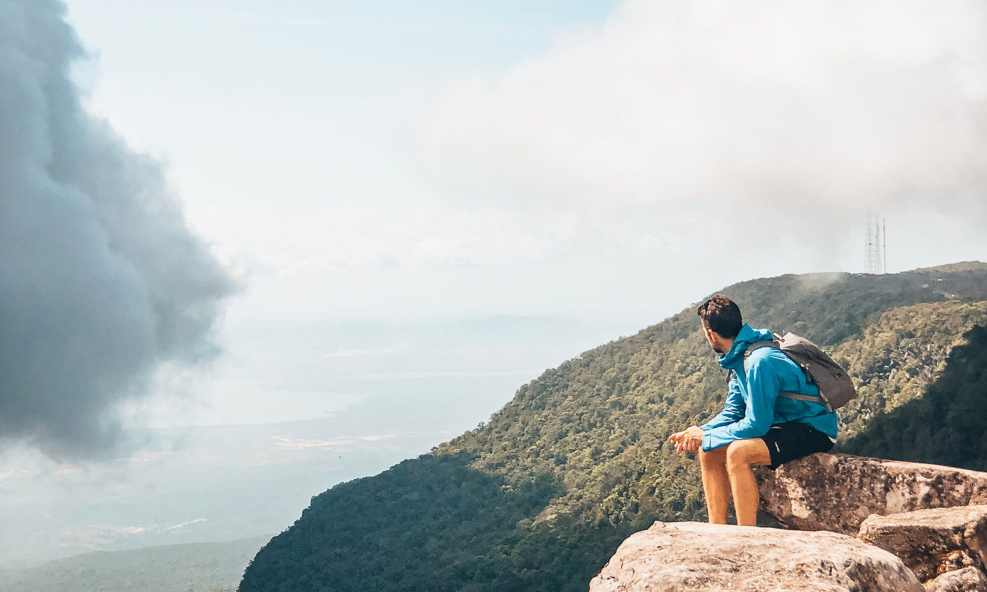 Man sits on rock overlooking hills and cloud on Bokor Mountain