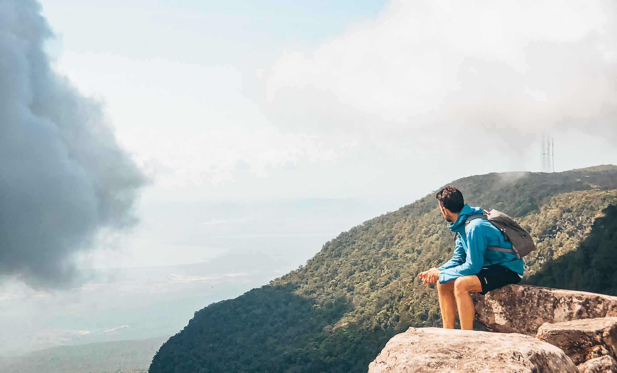 Man sits on rock overlooking hills and cloud on Bokor Mountain